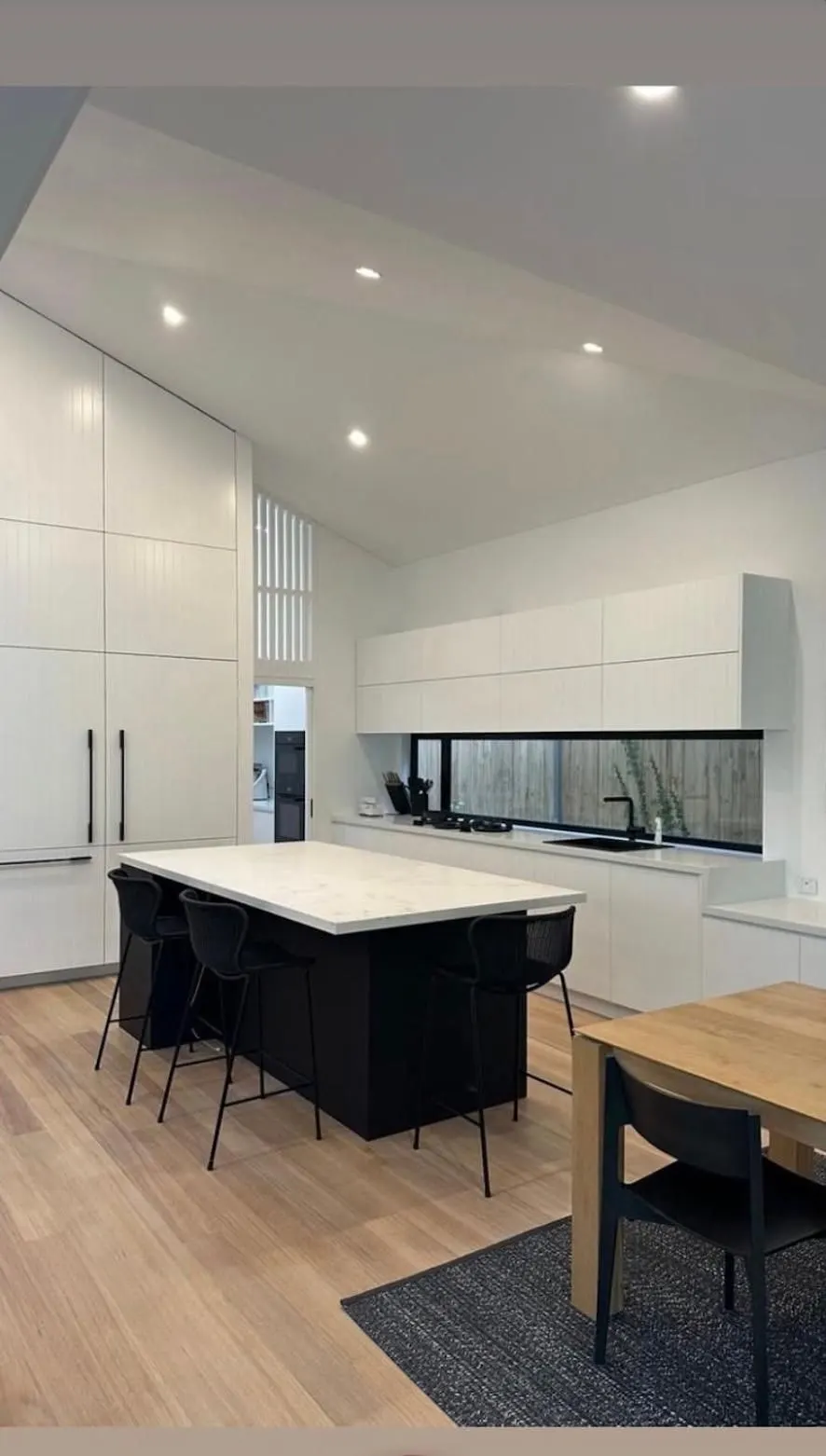 Kitchen with a black island bench, white floor to ceiling cabinetry, window splashback and vaulted ceiling