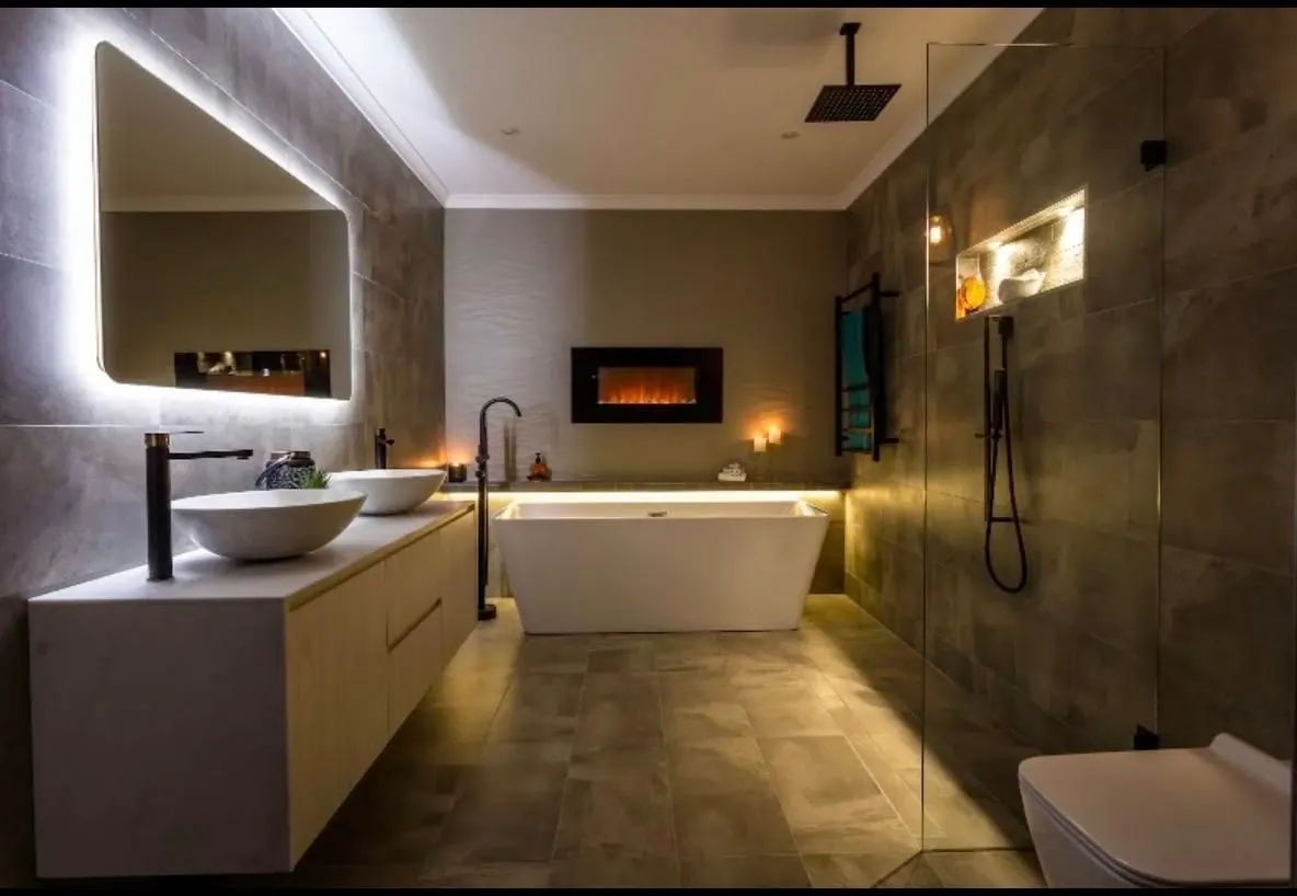 Luxury bathroom renovation with a freestanding bath, wall fireplace, backlit mirror and warm feature lighting