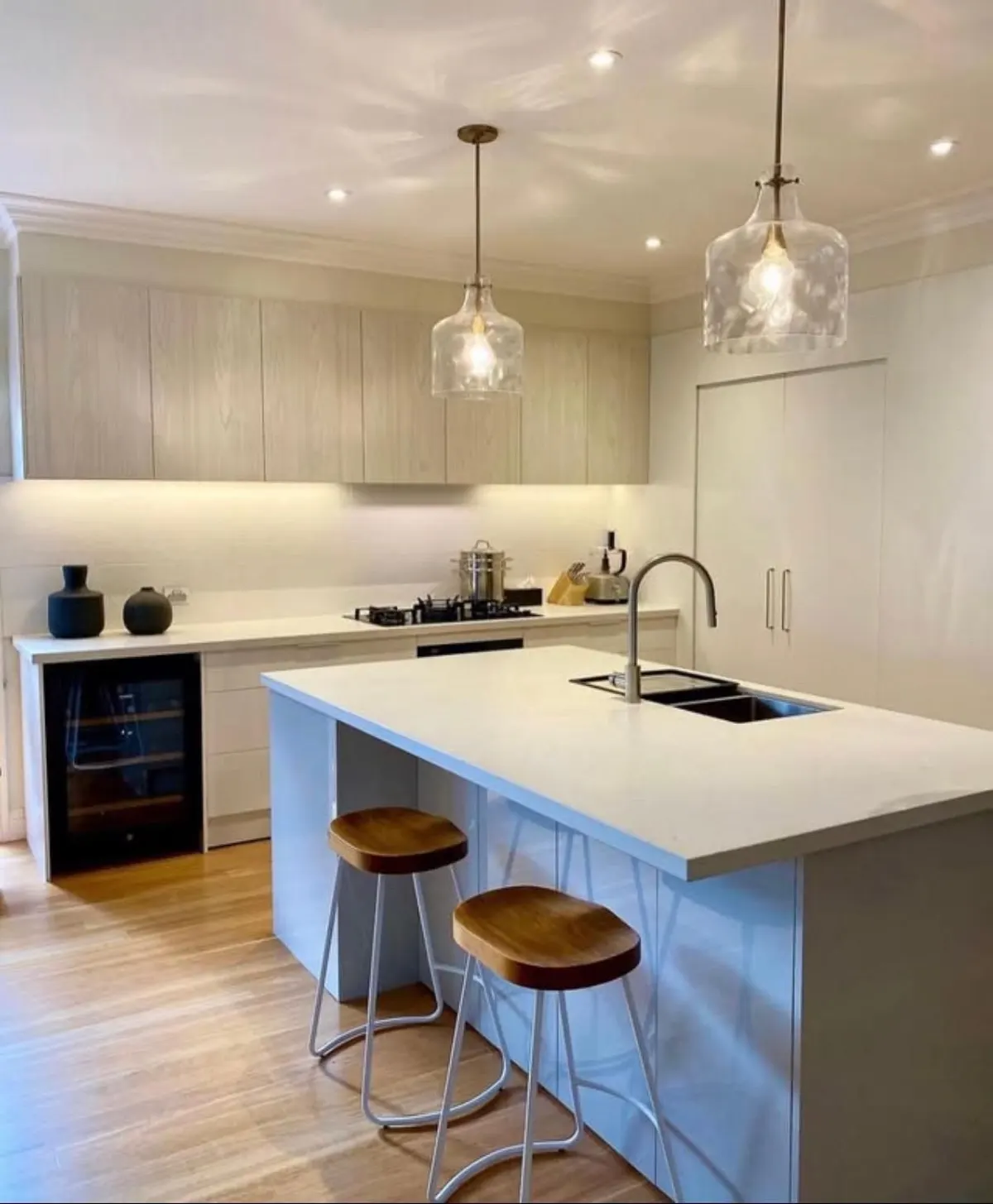 Kitchen renovation with a white island bench, glass pendant lights, timber stools, wine fridge and timber floors